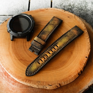 Puede incluir: Dos correas de reloj de cuero marrón con un acabado desgastado. Las correas tienen un patrón de color amarillo y marrón. Las correas están sobre una superficie de madera.