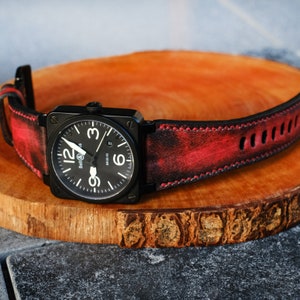 May include: A black square wristwatch with a black face and white numerals, the watch is on a red and black leather band. The watch is on a wooden surface.