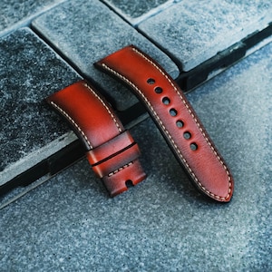 Pode incluir: Duas pulseiras de relógio de couro vermelho com costuras brancas. As pulseiras estão dispostas sobre uma superfície de pedra cinza.