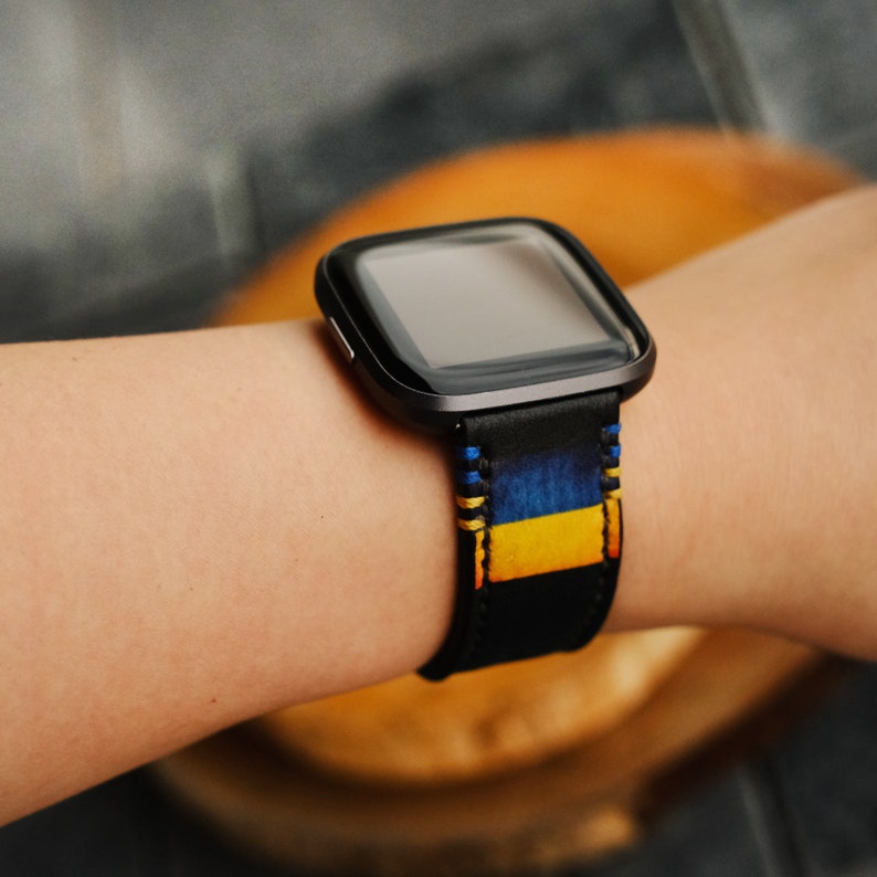 Puede incluir: Un reloj inteligente negro con una banda estampada en azul, amarillo y negro. La esfera del reloj es negra y tiene una pantalla digital.