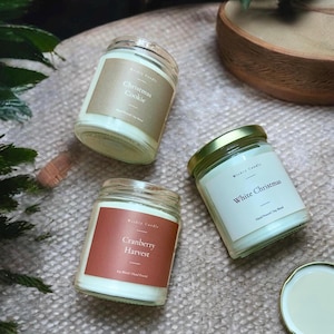 May include: Three glass jar candles with labels reading "Christmas Cookie", "Cranberry Harvest", and "White Christmas". The candles have white wax and are displayed on a neutral-toned surface. The jars have metal lids and are labeled "Weekly Candle".