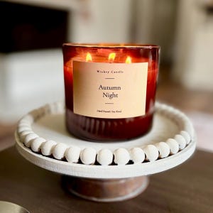 K&ouml;nnte beinhalten: Eine Drei-Docht-Kerze in einem dunkelroten Glasgef&auml;&szlig; mit einem beigen Etikett mit der Aufschrift Wickry Candle Autumn Night Hand Poured Soy Blend, platziert auf einem wei&szlig;en runden Fu&szlig;tablett mit einem Rand aus kleinen dekorativen Holzperlen.