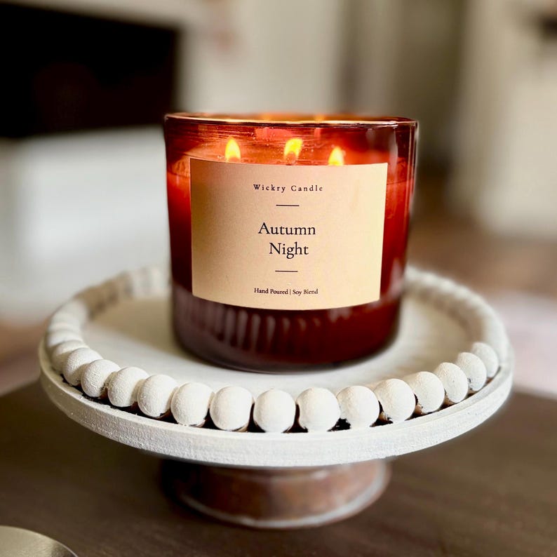 K&ouml;nnte beinhalten: Eine Drei-Docht-Kerze in einem dunkelroten Glasgef&auml;&szlig; mit einem beigen Etikett mit der Aufschrift Wickry Candle Autumn Night Hand Poured Soy Blend, platziert auf einem wei&szlig;en runden Fu&szlig;tablett mit einem Rand aus kleinen dekorativen Holzperlen.