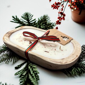 Op de afbeelding: Een ovale kaars in een houten kom gevuld met witte was, voorzien van een rood, groen en goud gestreept lint gestrikt in een strik. Een bruin papieren label met een uitgesneden kerstboomvorm zit aan het lint op het oppervlak van de kaars.
