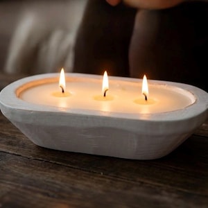 Op de afbeelding: Een ovale kaars in een witte houten kom met drie brandende lonten. De kaarswas is gebroken wit en vult de binnenkant van de uitgesneden houten houder. De buitenkant van de kom heeft een getextureerde, matte witte afwerking met zichtbare houtnerf.