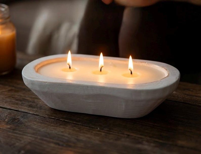 Op de afbeelding: Een ovale kaars in een witte houten kom met drie brandende lonten. De kaarswas is gebroken wit en vult de binnenkant van de uitgesneden houten houder. De buitenkant van de kom heeft een getextureerde, matte witte afwerking met zichtbare houtnerf.