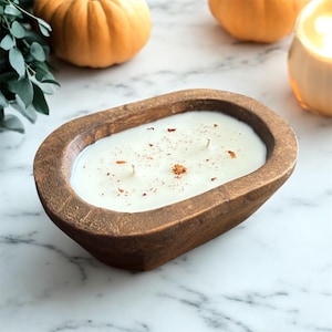 Op de afbeelding: Een witte waskaars met drie lonten in een ovale, donkerbruine houten kom. Het oppervlak van de was is bestrooid met kleine, gedroogde oranje botanische stukjes. De kom heeft een getextureerde afwerking met een natuurlijke houtnerf.