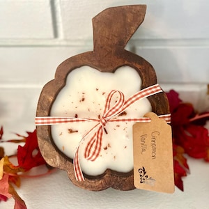 May include: A white candle in a dark brown wooden bowl shaped like an apple. The candle is topped with small brown spice flecks and tied with a red and white gingham ribbon. A tan paper tag attached to the ribbon reads Cinnamon Vanilla.