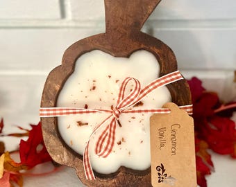 May include: A white candle in a dark brown wooden bowl shaped like an apple. The candle is topped with small brown spice flecks and tied with a red and white gingham ribbon. A tan paper tag attached to the ribbon reads Cinnamon Vanilla.
