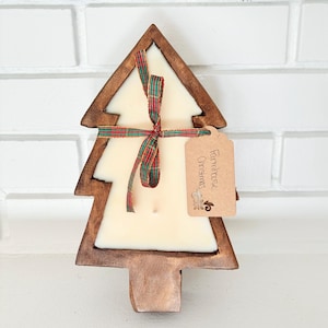 May include: A wooden Christmas tree candle holder with a white candle inside. The candle is tied with a red and green plaid ribbon. A brown tag is attached to the holder with the text "Farmhouse Christmas".