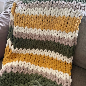 May include: A chunky knit throw blanket featuring horizontal stripes in alternating colors of olive green, mustard yellow, cream, and light taupe. The blanket is crafted from thick, soft yarn with a visible, large-scale looped stitch pattern throughout.