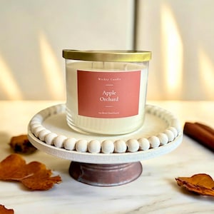 K&ouml;nnte beinhalten: Eine Kerze im klaren Glas mit goldenem Deckel und einem terrakottafarbenen Etikett mit der Aufschrift Wicky Candle Apple Orchard Soy Blend Hand Poured, platziert auf einem wei&szlig;en h&ouml;lzernen Tortenst&auml;nder mit perlenartigem Rand.