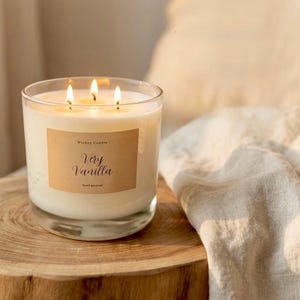 May include: A lit, hand-poured vanilla candle in a clear glass jar. The candle has three wicks and a label that reads "Very Vanilla." The candle sits on a wooden surface, with a neutral-colored cloth in the background.