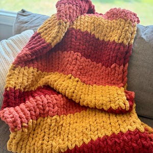 May include: A chunky knit throw blanket featuring thick, soft yarn in horizontal stripes of deep red, burnt orange, and golden yellow. The blanket has a highly textured, looped appearance created by large-scale knitting stitches throughout the fabric.