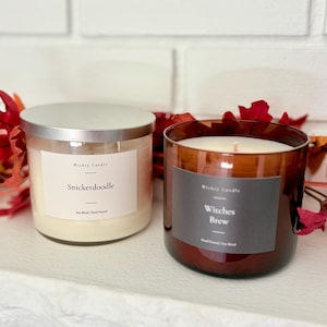 K&ouml;nnte beinhalten: Zwei Kerzen im Glas mit silbernen Deckeln. Die linke Kerze hat ein wei&szlig;es Etikett mit der Aufschrift Wickry Candle Snickerdoodle Soy Blend Hand Poured. Die rechte Kerze befindet sich in einem braunen Glas mit einem dunkelgrauen Etikett.