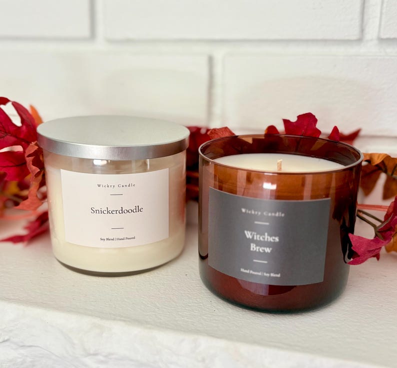 K&ouml;nnte beinhalten: Zwei Kerzen im Glas mit silbernen Deckeln. Die linke Kerze hat ein wei&szlig;es Etikett mit der Aufschrift Wickry Candle Snickerdoodle Soy Blend Hand Poured. Die rechte Kerze befindet sich in einem braunen Glas mit einem dunkelgrauen Etikett.