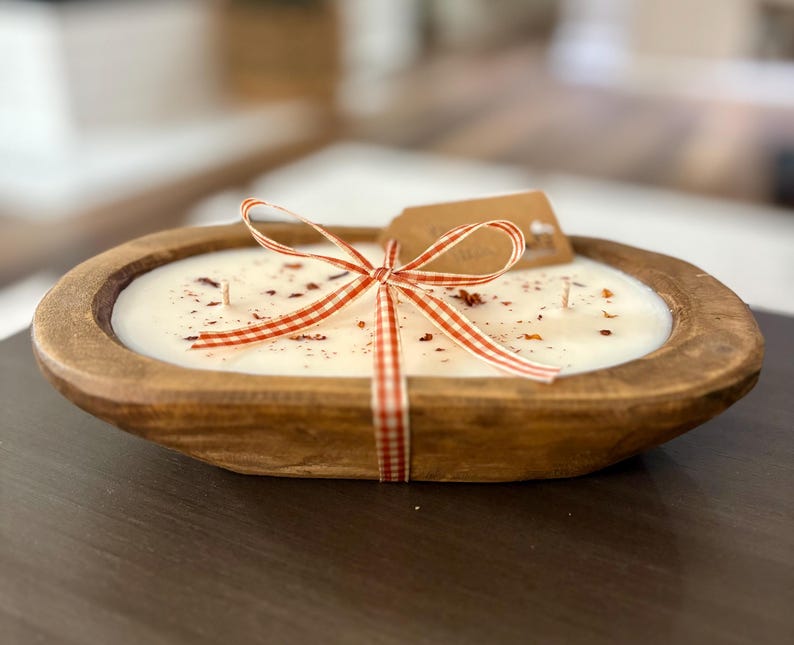 Op de afbeelding: Een ovale kaars in een houten deegkom gevuld met witte was en drie lonten. Het oppervlak van de was is bestrooid met gedroogde botanische stukjes. Een rood-wit geblokt lint is om de kom gestrikt, voorzien van een klein bruin label.