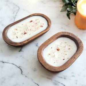 Op de afbeelding: Twee ovale houten deegkommen gevuld met witte was en elk drie lonten. Het oppervlak van de witte was is bestrooid met kleine, gedroogde roodbruine botanische stukjes. De kommen hebben een donkerbruine, getextureerde houtnerfafwerking.