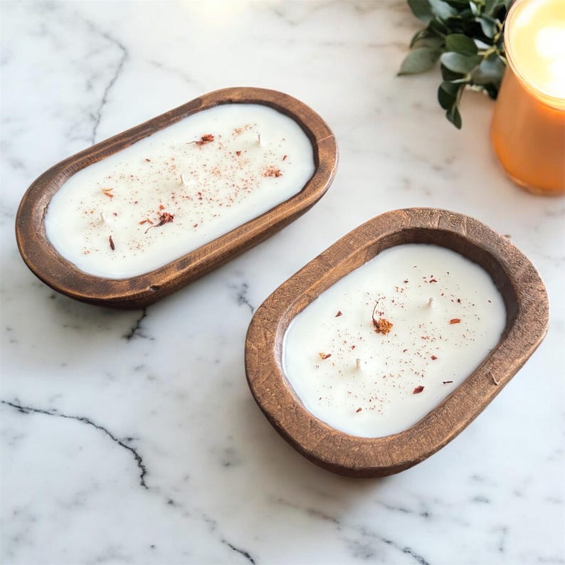 Op de afbeelding: Twee ovale houten deegkommen gevuld met witte was en elk drie lonten. Het oppervlak van de witte was is bestrooid met kleine, gedroogde roodbruine botanische stukjes. De kommen hebben een donkerbruine, getextureerde houtnerfafwerking.