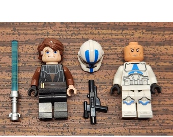 May include: Two Lego minifigures, one with brown hair and a black and brown outfit, the other with a white and blue outfit. The minifigure with brown hair is holding a blue lightsaber. The other minifigure is holding a black blaster. There is also a white and blue helmet.