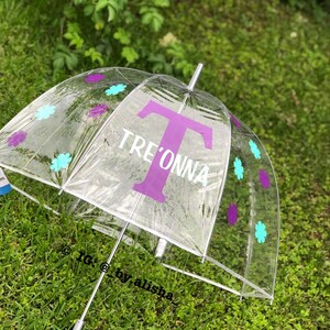 Op de afbeelding: Een transparante bubbelparaplu met een paarse letter "T" en de naam "Tre'onna" in wit. De paraplu is versierd met paarse en turquoise bloemen.