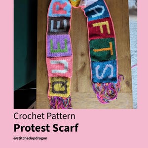 May include: A vibrant, hand-crocheted scarf displaying the words "REUSE CRAFTS" in bold, colourful letters. The scarf incorporates shades of pink, blue, green, and yellow, and has fringed edges. The text "Crochet Pattern Protest Scarf" is also visible.