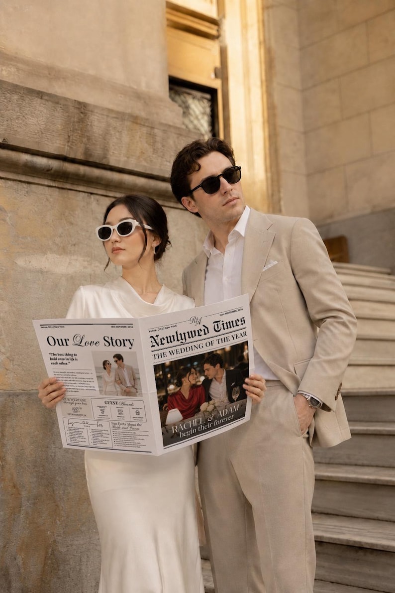May include: A couple in sunglasses poses with a newspaper titled "Newlywed Times" and "Our Love Story." The newspaper features wedding photos and the headline "The Wedding of the Year." The man wears a tan suit, and the woman wears a white dress.