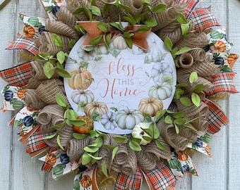 May include: A fall-themed wreath with a circular center featuring pumpkins and the text "bless this home." The wreath is adorned with burlap, orange plaid ribbons, and green foliage, creating a festive autumn decoration.