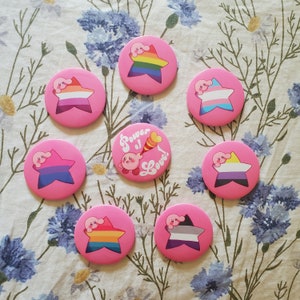 May include: Ten pink and white button pins featuring a pink character with a star and the text "Power of Love!" on one pin. The other pins feature the character in different poses with a rainbow, a heart, and a star.