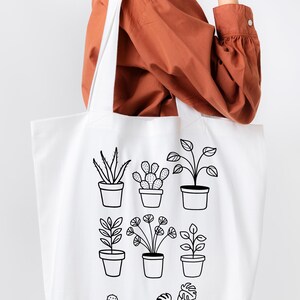 May include: White canvas tote bag featuring black line drawings of potted plants. The bag has the text "PIPPA & PINE" printed on the bottom.