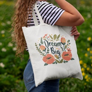 May include: A white tote bag with a floral design and the text "Dream Big". The bag has two long straps and is being held by a person.
