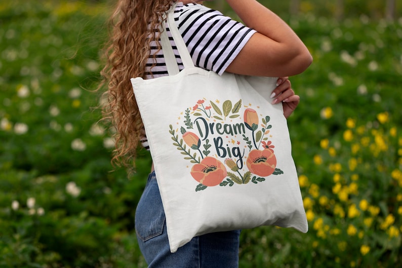 May include: A white tote bag with a floral design and the text "Dream Big". The bag has two long straps and is being held by a person.