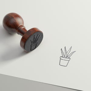 May include: A wooden stamp with a black rubber surface featuring a line drawing of a potted plant. The stamp is on a white surface with the text "PIPPA & PINE" in the bottom right corner.
