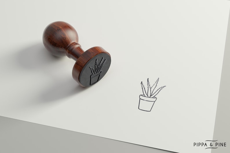 May include: A wooden stamp with a black rubber surface featuring a line drawing of a potted plant. The stamp is on a white surface with the text "PIPPA & PINE" in the bottom right corner.