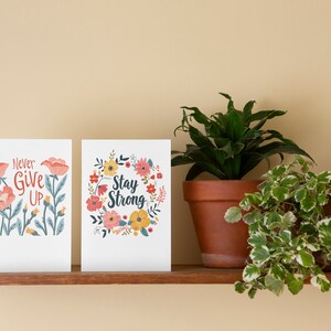May include: Two floral-themed cards with inspirational messages. The left card says "Never Give Up" and the right card says "Stay Strong". The cards are on a wooden shelf with two potted plants.