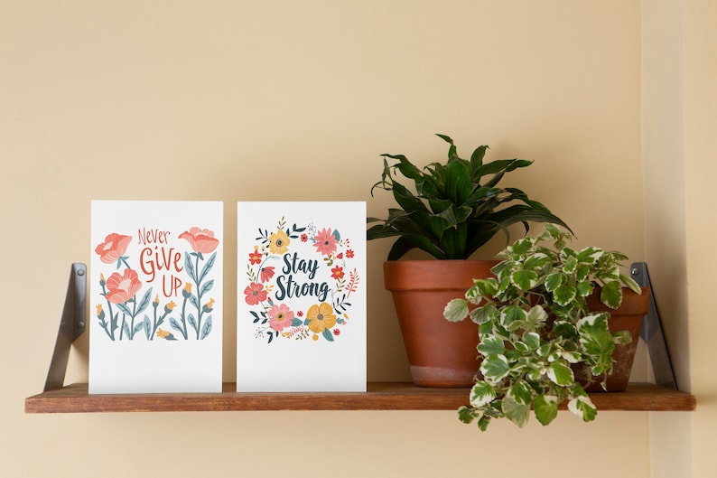 May include: Two floral-themed cards with inspirational messages. The left card says "Never Give Up" and the right card says "Stay Strong". The cards are on a wooden shelf with two potted plants.