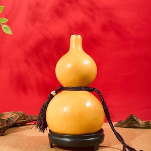 Peut inclure: Un récipient lisse en forme de double gourde jaune clair, doté d'un col étroit et d'une base large. La gourde repose sur un support noir circulaire et est ornée d'un cordon tressé marron foncé avec un pompon et une petite perle décorative.