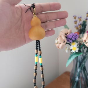 May include: A gourd-shaped ornament with a gold-coloured metal loop and black string. The ornament has a beaded strand with black, yellow, and brown beads. A vase of flowers is in the background.