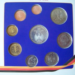 Op de afbeelding: Een verzameling van tien Duitse Deutsche Mark-munten in een blauwe en gouden display case. De munten zijn in een cirkel gerangschikt met de 10 Deutsche Mark-munt in het midden. De munten zijn allemaal zilver en goud van kleur.