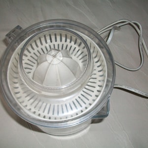 Op de afbeelding: Een witte elektrische citruspers met een doorzichtig plastic deksel en een wit pulpfilter. De citruspers heeft een snoer en een stekker.