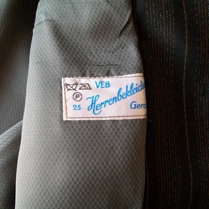 May include: A close-up of a gray fabric with a white label that reads "VEB Herrenbekleidung Gera 25". The label has a blue border and includes a washing instruction symbol.