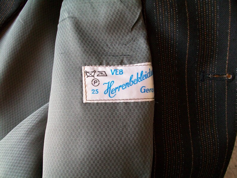 May include: A close-up of a gray fabric with a white label that reads "VEB Herrenbekleidung Gera 25". The label has a blue border and includes a washing instruction symbol.