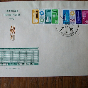 May include: A white envelope with a gold logo and the text "LEIPZIGER HERBSTMESSE 1973". The envelope has a series of stamps with images of buildings, a guitar, and a bowling pin. The stamps are from the German Democratic Republic (GDR).