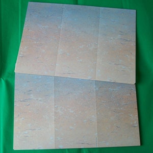May include: Two unfolded, rectangular cardboard sheets with a mottled, stone-like pattern in shades of beige, tan, and blue. The sheets are divided into sections by vertical creases. The background is a solid green.