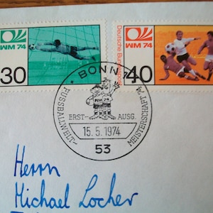 Könnte beinhalten: Zwei deutsche Briefmarken aus dem Jahr 1974 mit Fussballmotiven. Die Briefmarken sind grün und orange mit dem Text "WM 74" und "Deutsche Bundespost". Ein runder Poststempel lautet "Fussballwelt- Meisterschaft 74" mit dem Datum "15.5.1974" und der Nummer "53".