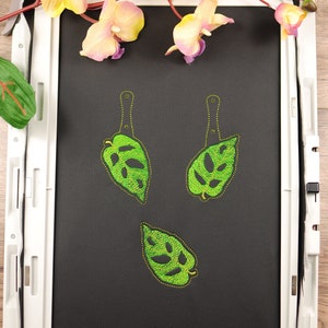 May include: Three green monstera leaf embroidery designs on a black fabric in an embroidery hoop.