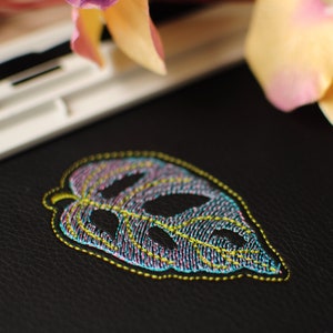 May include: A close-up of a black leather surface with a colorful embroidered leaf design. The leaf is outlined in yellow thread and filled with purple, blue, and green thread.
