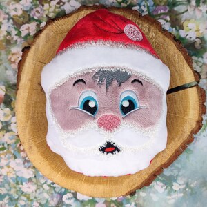 May include: Embroidered Santa Claus face with a red hat, white beard, and blue eyes. The face is on a wooden background.