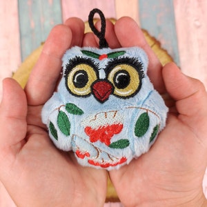 May include: A blue plush owl ornament with embroidered details, including red berries, green leaves, and black eyes.