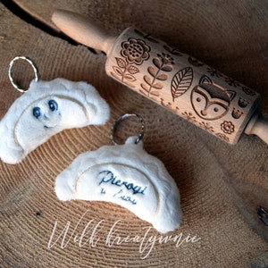 Pode incluir: Dois chaveiros de pierogi de pelúcia branca com rostos bordados e o texto "Pierogi 4 Life" em um. Também é mostrado um rolo de massa de madeira com um design de raposa e flores.